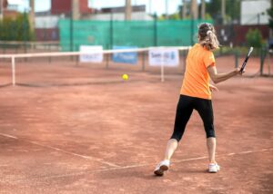 Woman playing tennis on clay court wearing orange sportswear, back view.